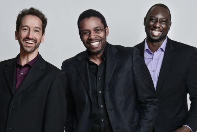 Three artists smile before a pale backdrop, wearing dark jackets and bright shirts.