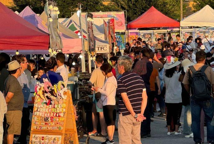 Busy outdoor market with colorful stalls, lights and crowds browsing items in a lively festival setting.