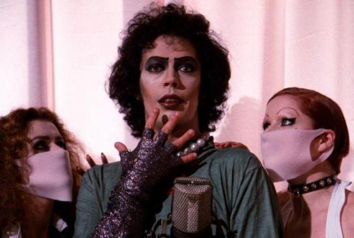 Three figures stand close together central with curly hair dark makeup and glove others with covered mouths and dark style.