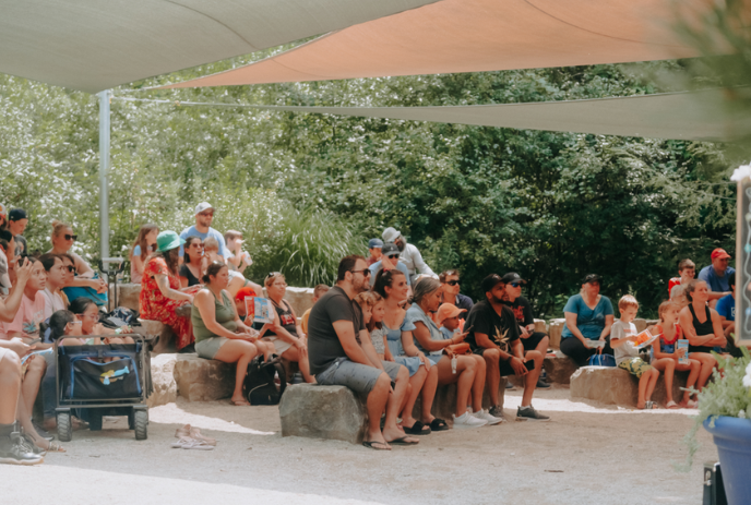 A group of people sit together in the shade watching a performance