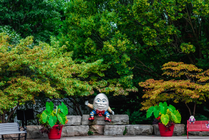 Humpty Dumpty sits on a wall waving, surrounded by green trees and plants.