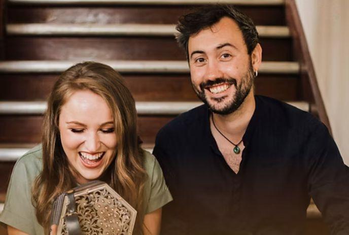 Caroline Keane and Tom Delaney sitting on stairs holding patterned object and paper tubes in a calm black and white scene.