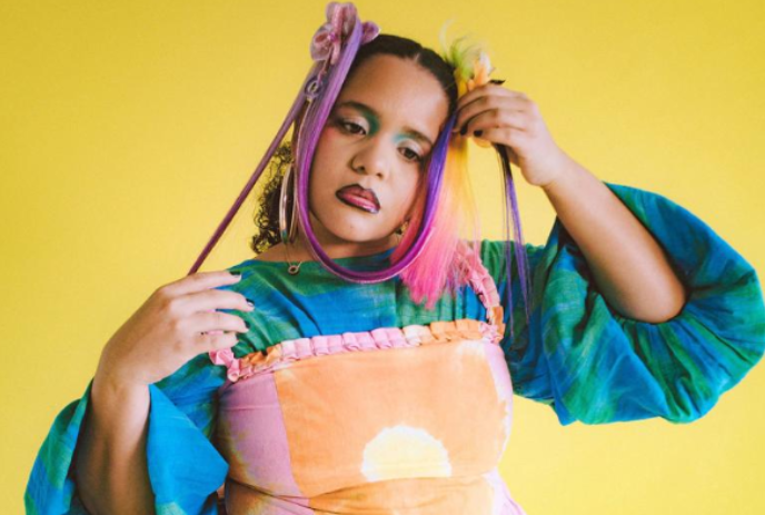 Person with colourful hair and a bright green and turquoise top in front of a yellow background.