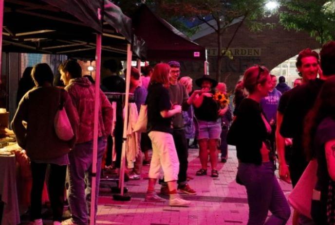 Evening market scene with tents vendors shoppers and bright pink purple lighting creating lively atmosphere.
