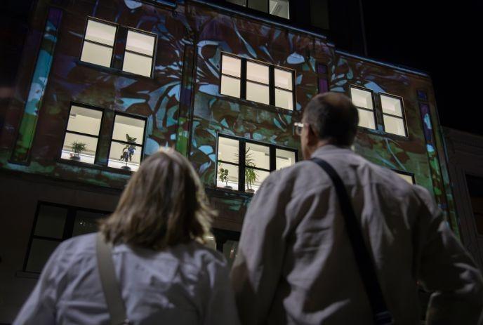 Two people stand outdoors at night watching colorful abstract light projections covering windows of tall building.