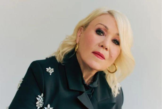 Portrait shot of Jann Arden with short blond hair in a black embroidered jacket, looking  forward.