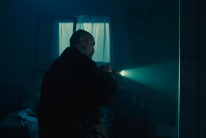 Person holding a flashlight in a dark room, looks around the wall at something not visible to the audience.