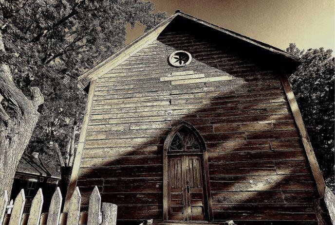 Old wooden chapel with arched doorway and steep roof surrounded by trees and white fence.