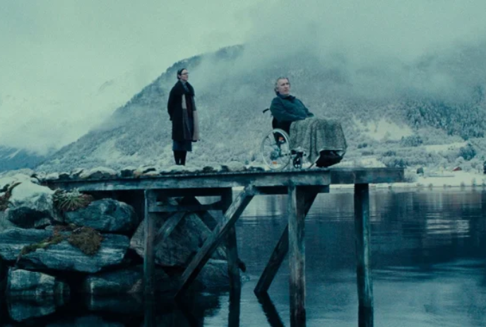A person in a wheelchair sits at the end of a dock with snowy mountains in the distance, while another person looks on.