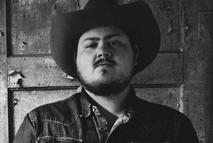Artist in cowboy hat and denim shirt standing before rustic weathered wall in black and white.