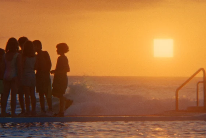 A group of young people gather around something while standing in front of a sunset beach area.