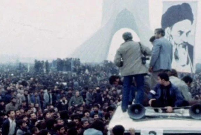 People standing on a vehicle in front of a large crowd with a leader's image beside them.