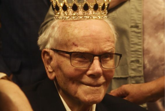 An older person sits with a crown on their head and people surrounding them.