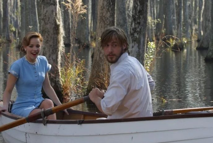 A man wearing a white shirt and a woman wearing a blue dress are in a rowboat surrounded by water and trees.