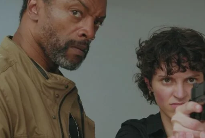 A man and a woman stand together while the woman aims a gun at something unseen.