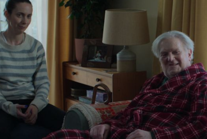 Two people sit together in a living room watching something off-screen.