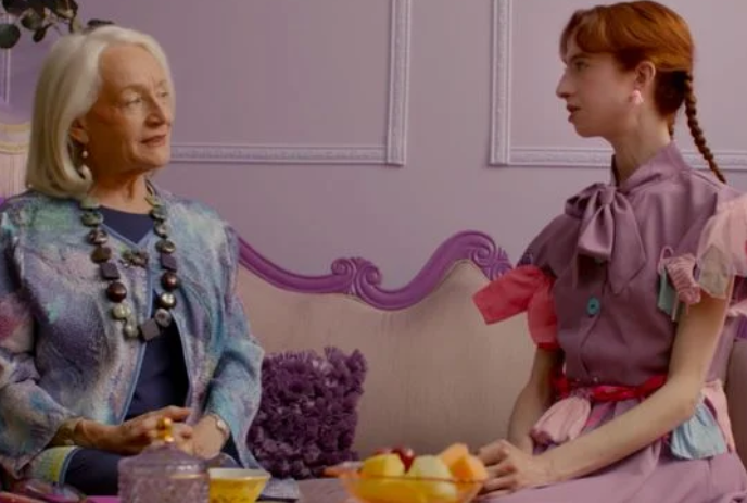A young woman in a purple dress sits upright with an older woman in a turquoise and purple jacket at a table with fruit on it.