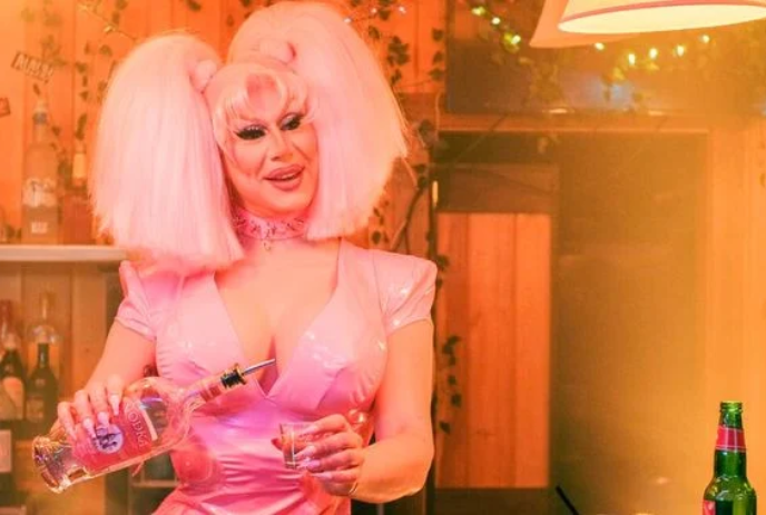 A person with extreme make-up, pink hair and a pink dress pours a pink drink into a shot glass.