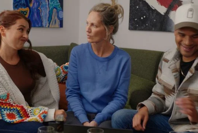 Three people sit together on a green corner sectional with paintings in the background.