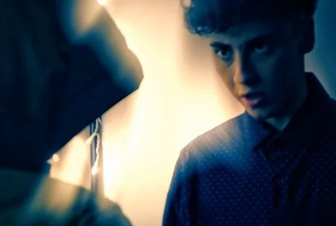 A young person stands intently in front of another person wearing a hoodie.
