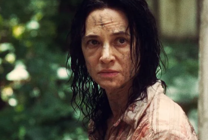 A woman with bedraggled hair and dirt on her face and clothing looks toward something off-screen.