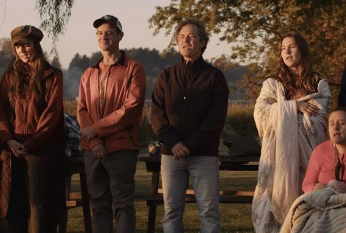 A group of people stand solemnly together in a park with trees, while singing.