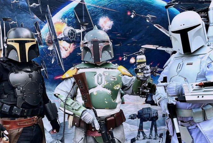 Armored trio in detailed Mandalorian cosplay posed before a vivid outer?space battle backdrop scene.