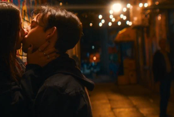 Two young people standing in an alleyway share a kiss, while someone else walks past them.