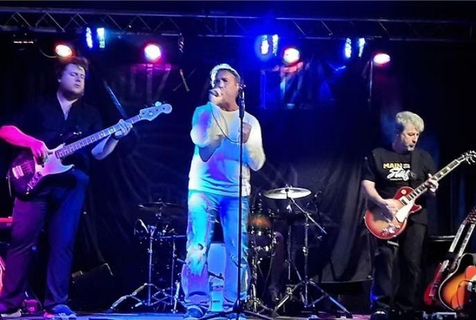 Three musicians performing live on a lit stage with guitars, drums and vibrant colored concert lighting.