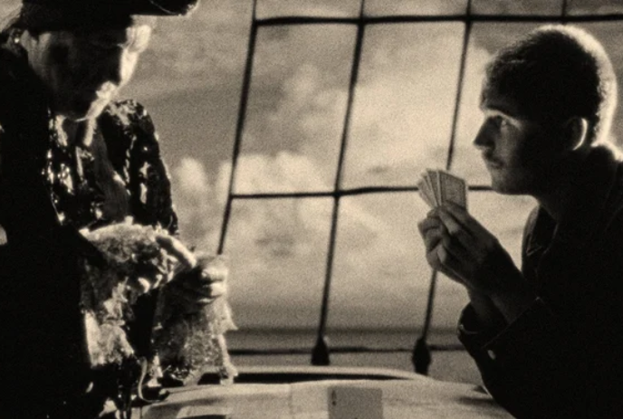 Black and white image of a person playing cards with what looks like a pirate out at sea.