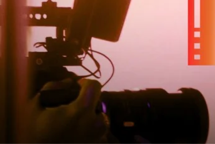 A person's hand holding an SLR camera with pink overtones in the background.