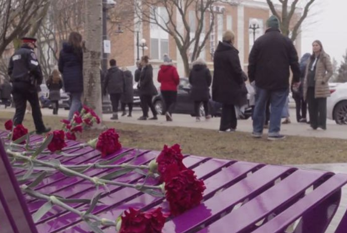 People mill around in front of a purple bench that has a number of red carnations on it.