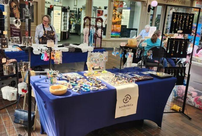 Jewelry vendor booth with blue tablecloth displaying handmade bracelets, earrings, and necklaces at indoor market.