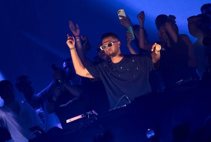Matt Sassari in white sunglasses raising hands on blue?lit stage as crowd cheers and captures the lively moment.