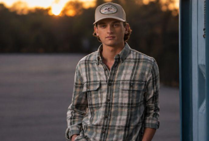 Person in plaid shirt and cap standing outdoors near a blue structure at sunset, hands in pockets.