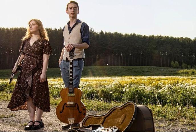 Two musicians with guitars standing in a flower-filled field beside an open guitar case.