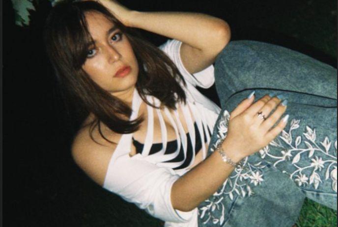 Artist Lelê seated outdoors wearing a cutout white top and embroidered jeans.