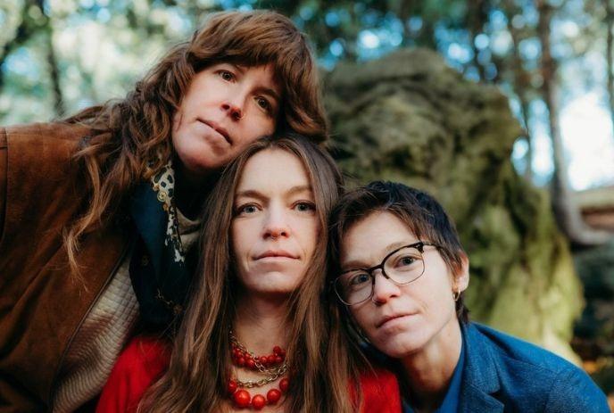 Three artists posing closely together, outdoors with trees and rocks in the background..