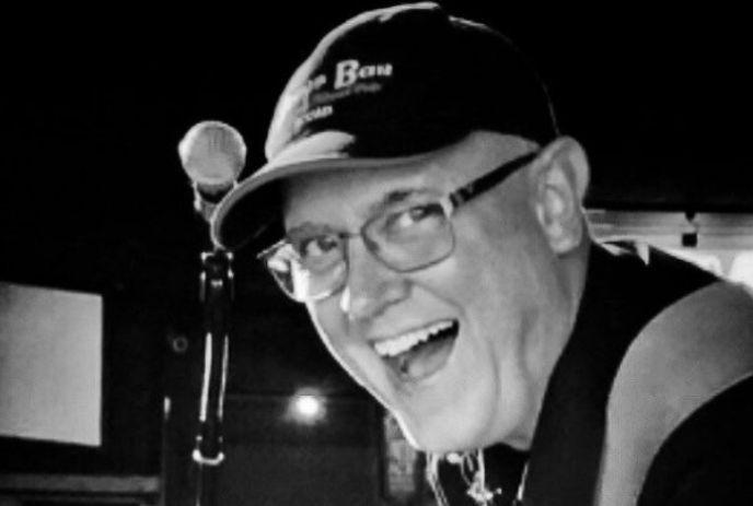 Black-and-white portrait of Pete Denomme in glasses and a cap beside a microphone stand.