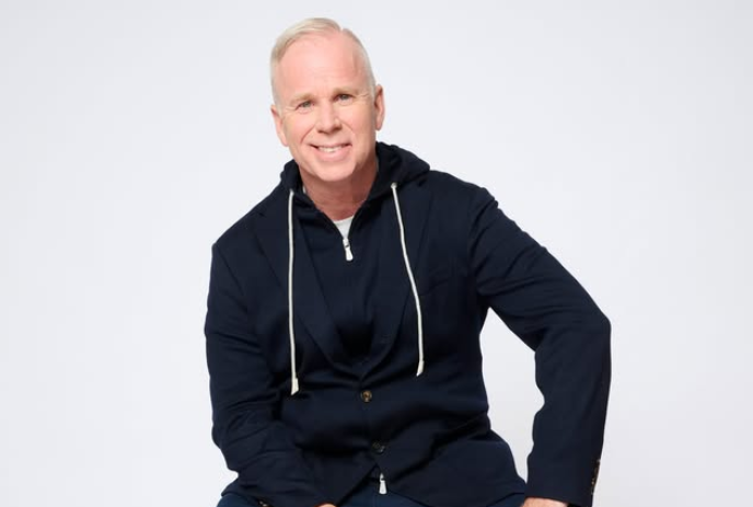 Gerry Dee sits wearing a blue hoodie under a blue sport coat and smiling at the camera.