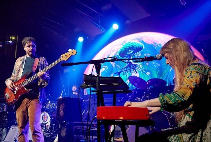 Live band performs on stage with keyboard, bass guitar, drums, and colorful psychedelic backdrop.