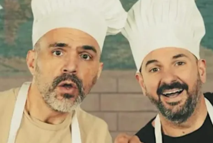 Two people wearing chef hats and aprons stand together looking forwards.