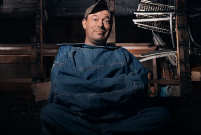 A person wearing a baseball cap and a backwards jean jacket sits in a garage surrounded by broken shelves.