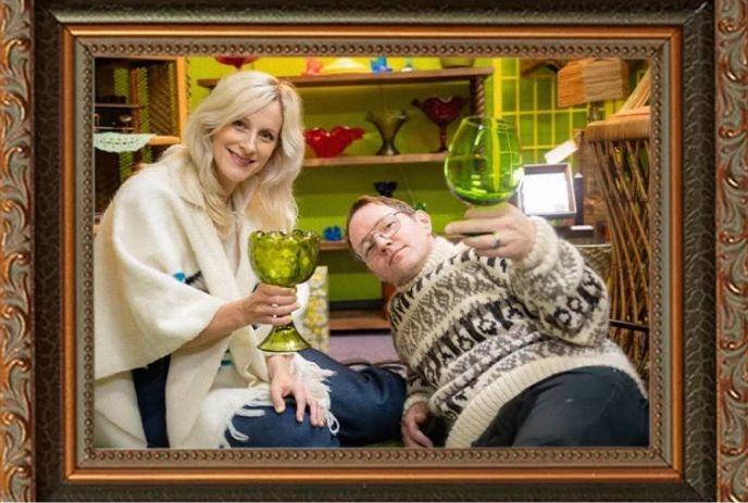 Two artists pose inside an ornate frame, each holding a green glass goblet in a colorful room.