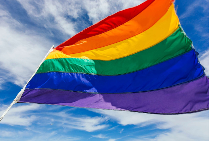 A vibrant rainbow flag with six colorful stripes waves proudly against a bright blue sky with scattered clouds.
