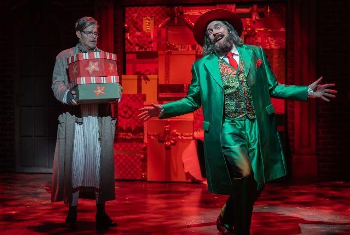 Festive actors on stage with gifts and a colorful green suited figure in a lively holiday setting