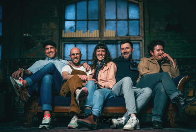 Five people sitting close together on a sofa in a dimly lit room with a dark window in the background.