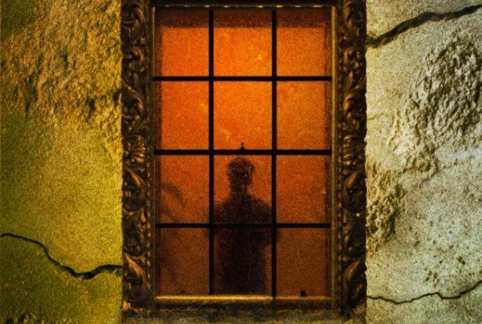 Silhouette of a person standing behind a warmly lit window set into a cracked textured wall.