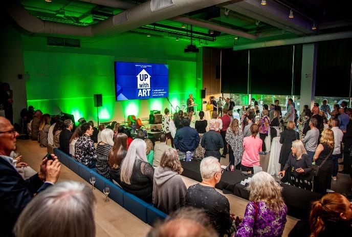 A crowd in a dimly lit event space watches a speaker on stage. A screen displays 'Up with Art.'