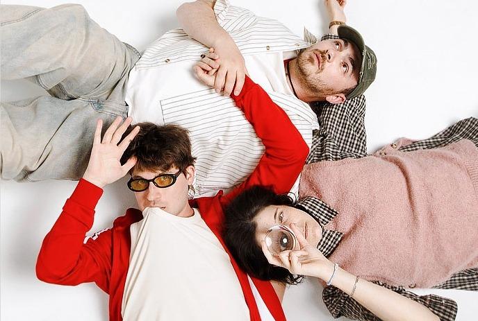 Three people lying together on a light surface, one holding a glass and one wearing sunglasses.